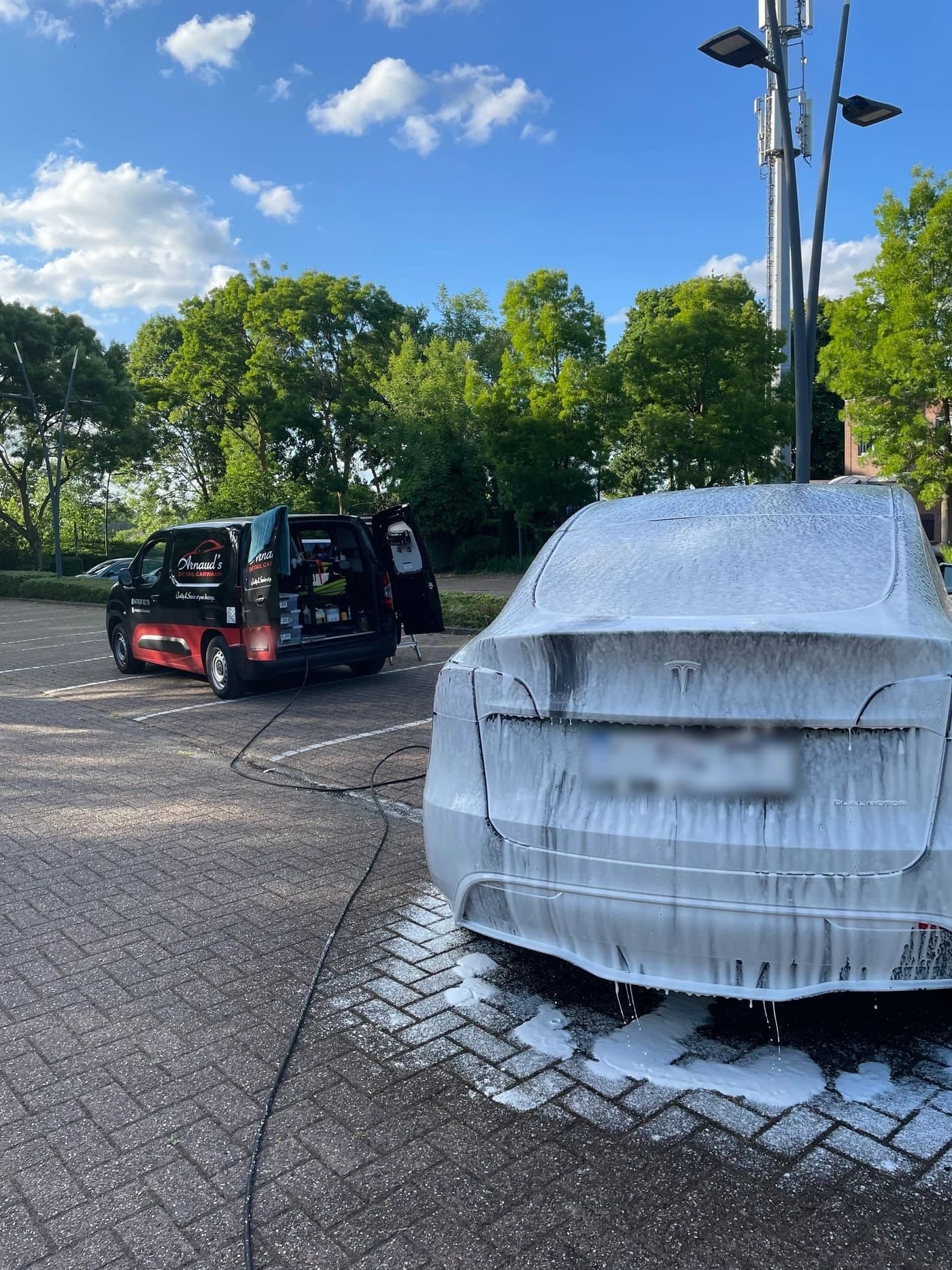 Een witte wagen onder het schuim tijdens een handwas, met de bestelwagen van Arnaud's Detail Carwash op de achtergrond